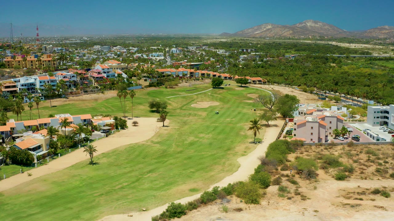 Aerial view over a San Jose Del Cabo golf course with green fairways, sand traps, residential buildings, palm trees, distant mountains, clear sky, and no visible people.