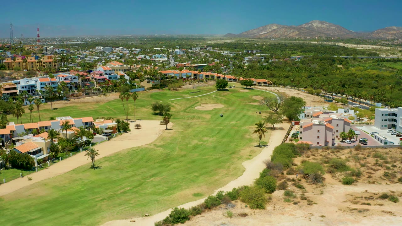 San Jose Del Cabo aerial frame showing broad green fairway, pale bunkers, orange-roofed homes, palms, dry lots, distant hills, and no visible people.