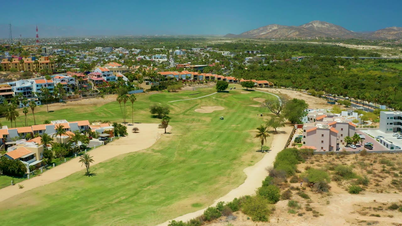 Wide San Jose Del Cabo aerial with manicured fairway in front, sandy bunkers, nearby residential blocks, palms, dense trees beyond, mountains, and no visible people.