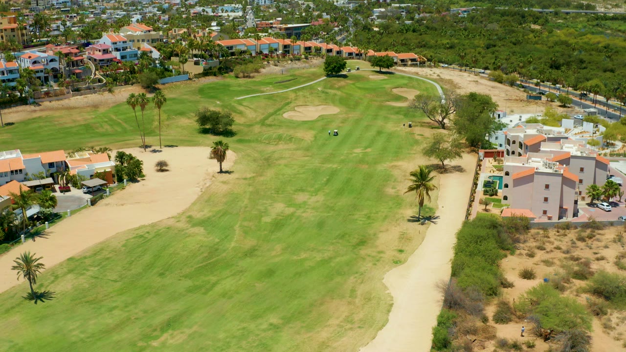 Aerial view over a San Jose Del Cabo golf course, showing green fairways, pale sand traps, palm trees, dry scrub, and red-roofed residential buildings at the course edge with no visible people.