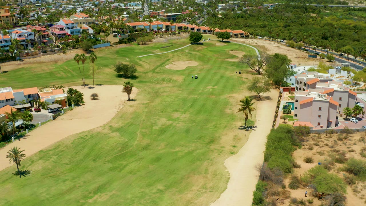 Overhead golf fairway bordered by sand paths, palms, and red-roofed buildings.