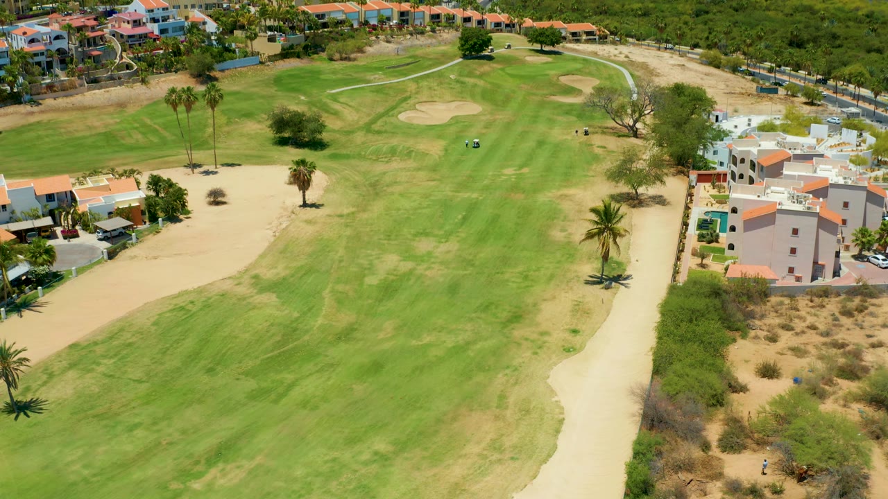 Aerial frame of the San Jose Del Cabo course with a broad green fairway, a sand trap near the upper right, palms, dry rough, and white residential buildings with red roofs along the right side with no visible people.