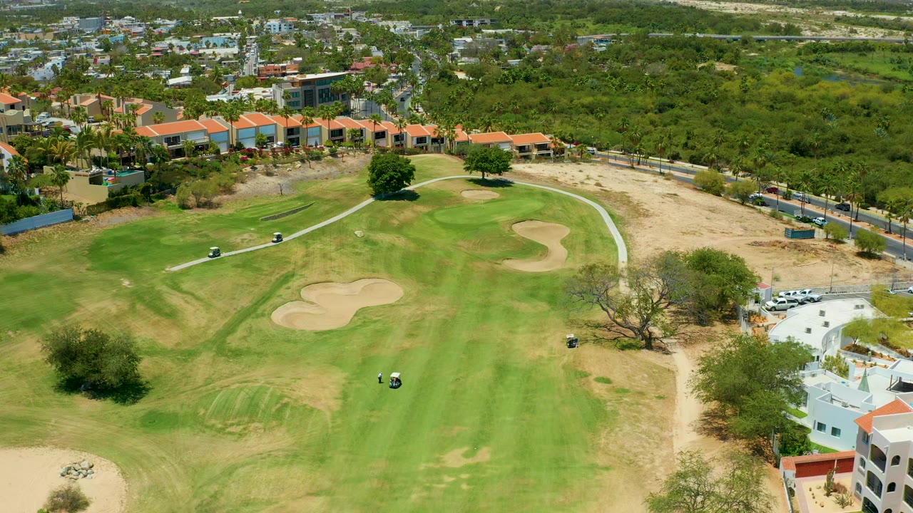 Drone view of golf carts and people on a green beside sand traps and villas.