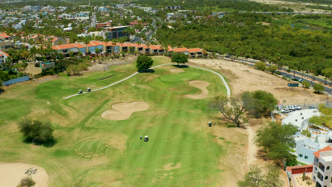 Aerial frame in San Jose Del Cabo showing a broad golf course green, two sand traps, a cart path, red-roofed buildings, dry ground edges, and no visible people.
