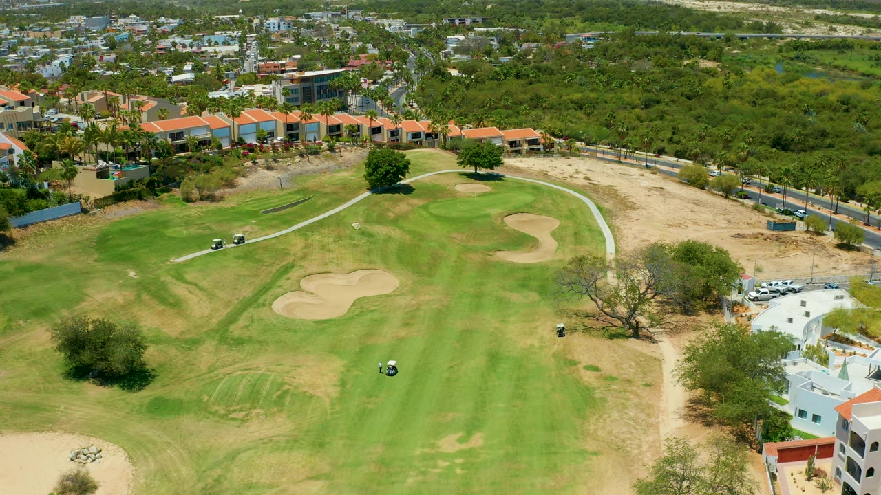 Aerial frame from San Jose Del Cabo showing a green golf fairway, tan sand traps, small carts, nearby white and terracotta-roofed buildings, trees, roads, and no visible people.