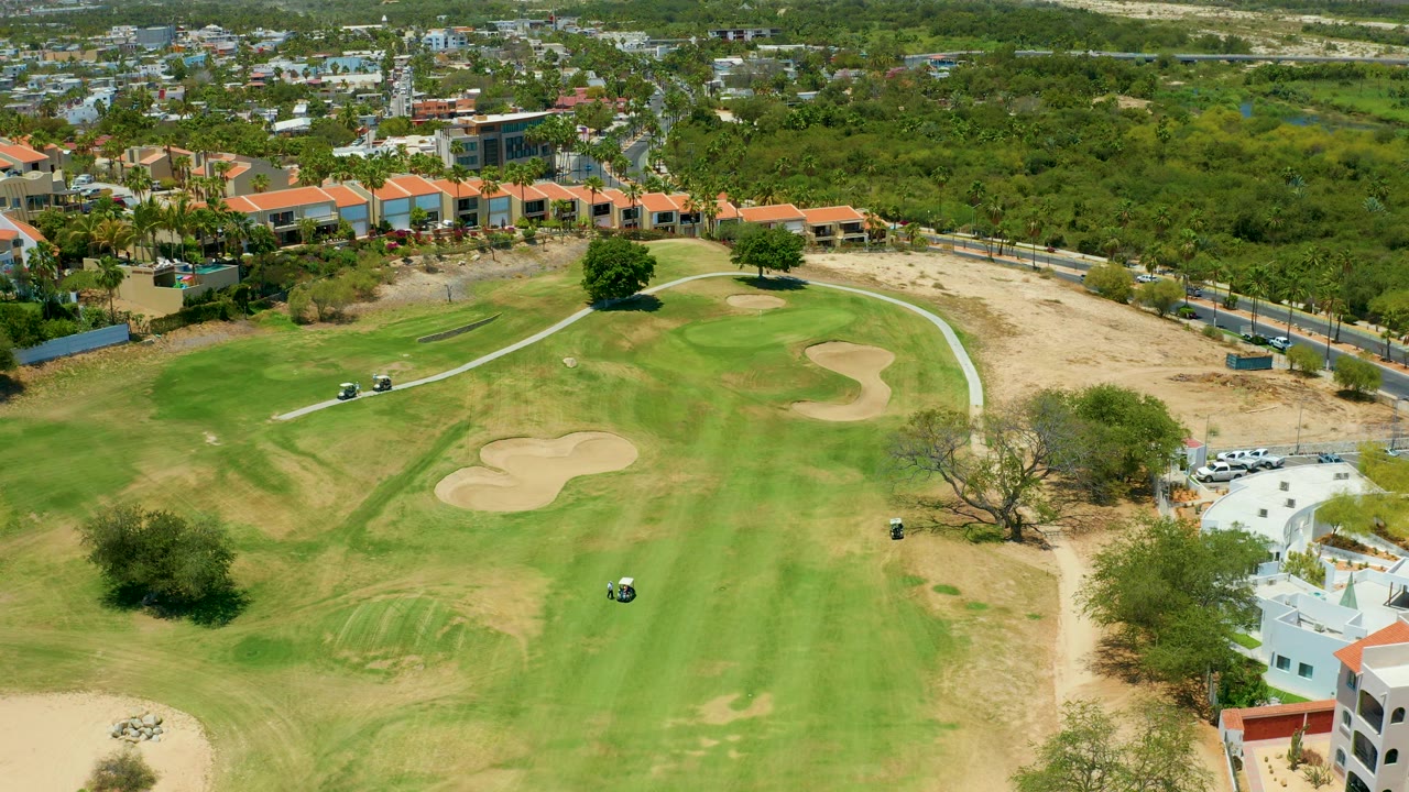 Aerial frame in San Jose Del Cabo looking over golf-course grass, sand traps, small carts, white buildings at the lower right edge, roads, dense trees, and no visible people.