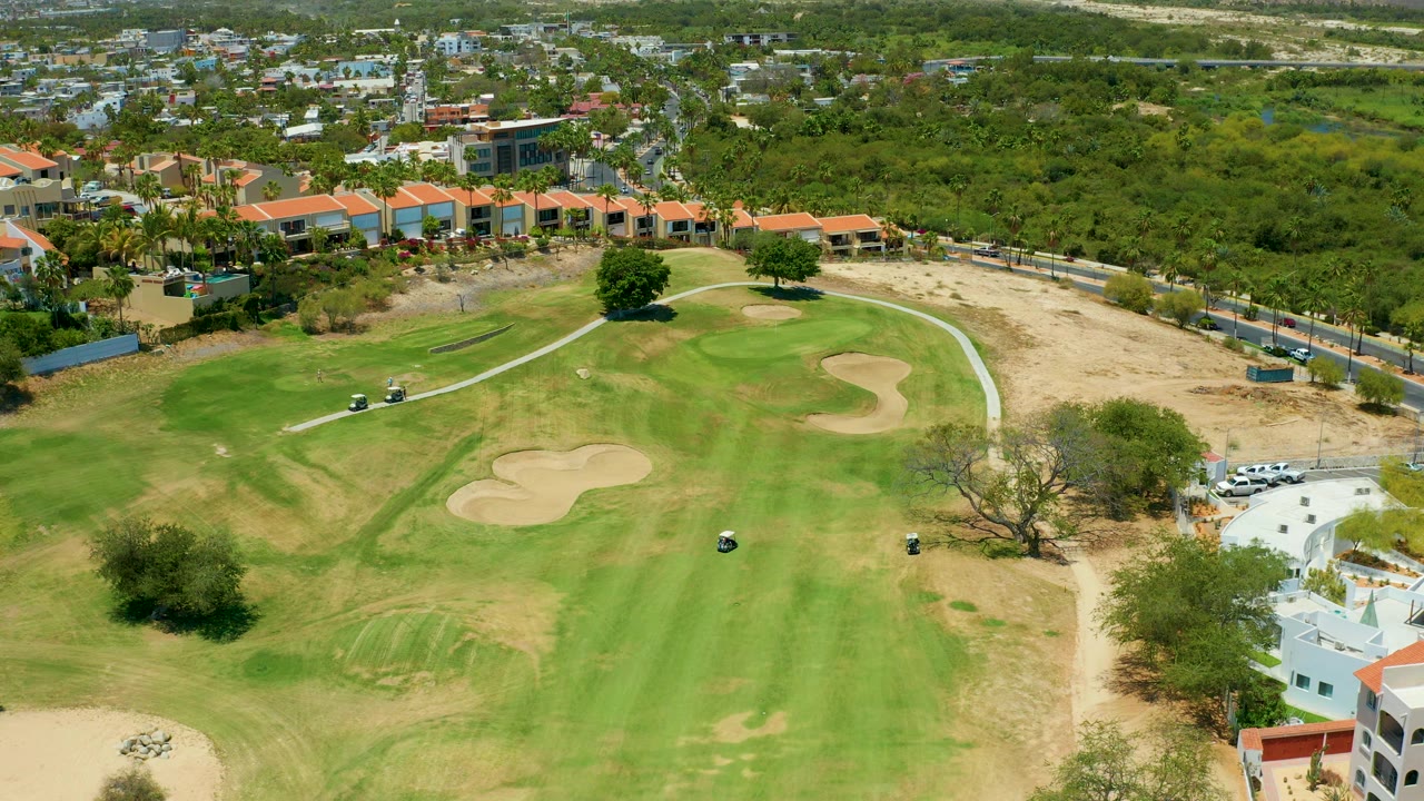 Aerial frame of the San Jose Del Cabo course with golf carts slightly farther along the path, sand traps on the fairway, trees, dry lots, and no visible people.
