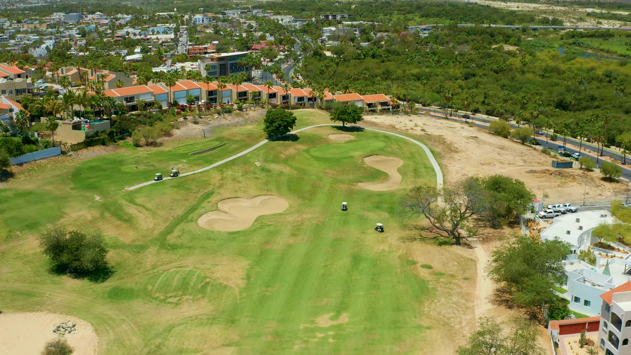 Opening aerial frame of a San Jose Del Cabo golf course with green fairways, tan sand traps, a few golf carts on the paths, low buildings in the background, and no visible people.