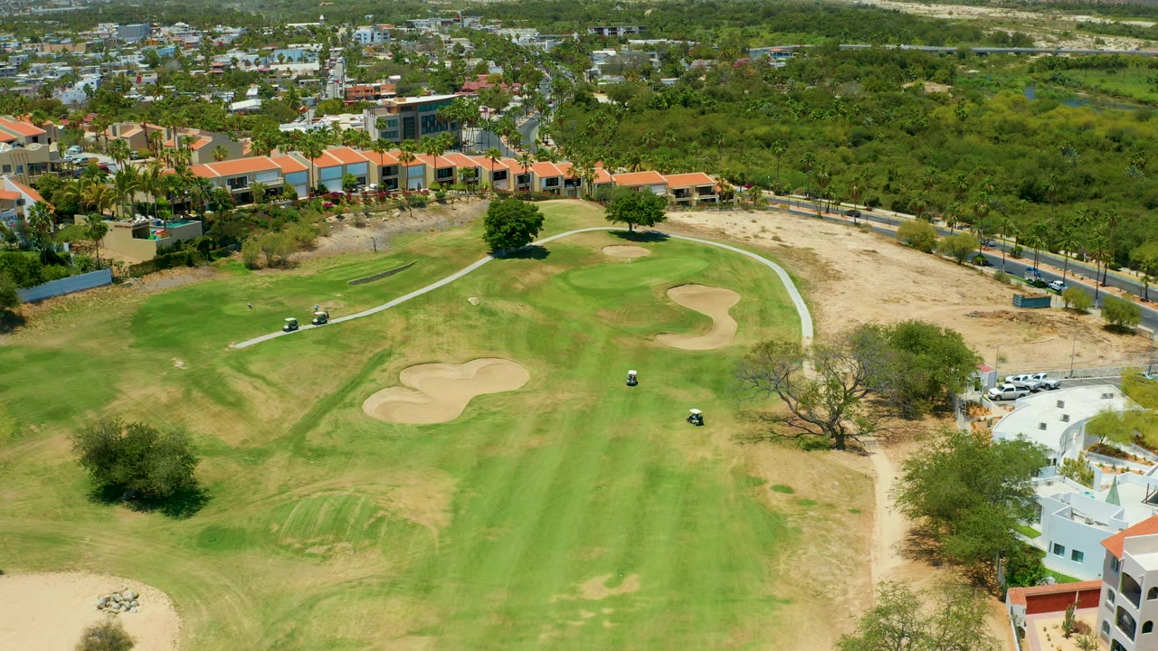 Elevated golf course view with carts near a curving path, sand traps, trees, and low buildings.