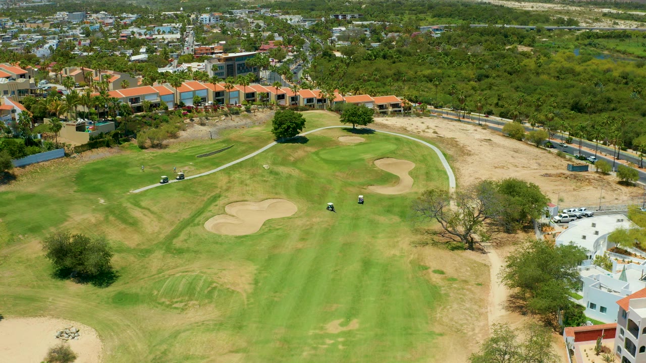 Aerial frame from San Jose del Cabo showing a green golf fairway, pale sand traps, a curving cart path with golf carts, nearby buildings, dry open land, and no visible people.