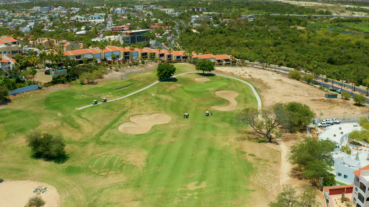 High aerial frame of the San Jose del Cabo golf course with the fairway centered, sand traps below the red-roofed buildings, golf carts near the path, dry ground at right, and no visible people.