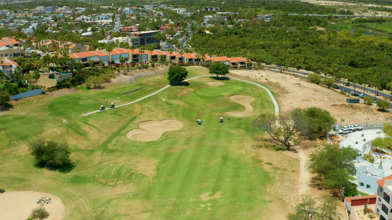 Aerial frame of a San Jose Del Cabo golf course showing green fairway, two pale sand traps, small golf carts on a curved path, buildings in the background, and no visible people.