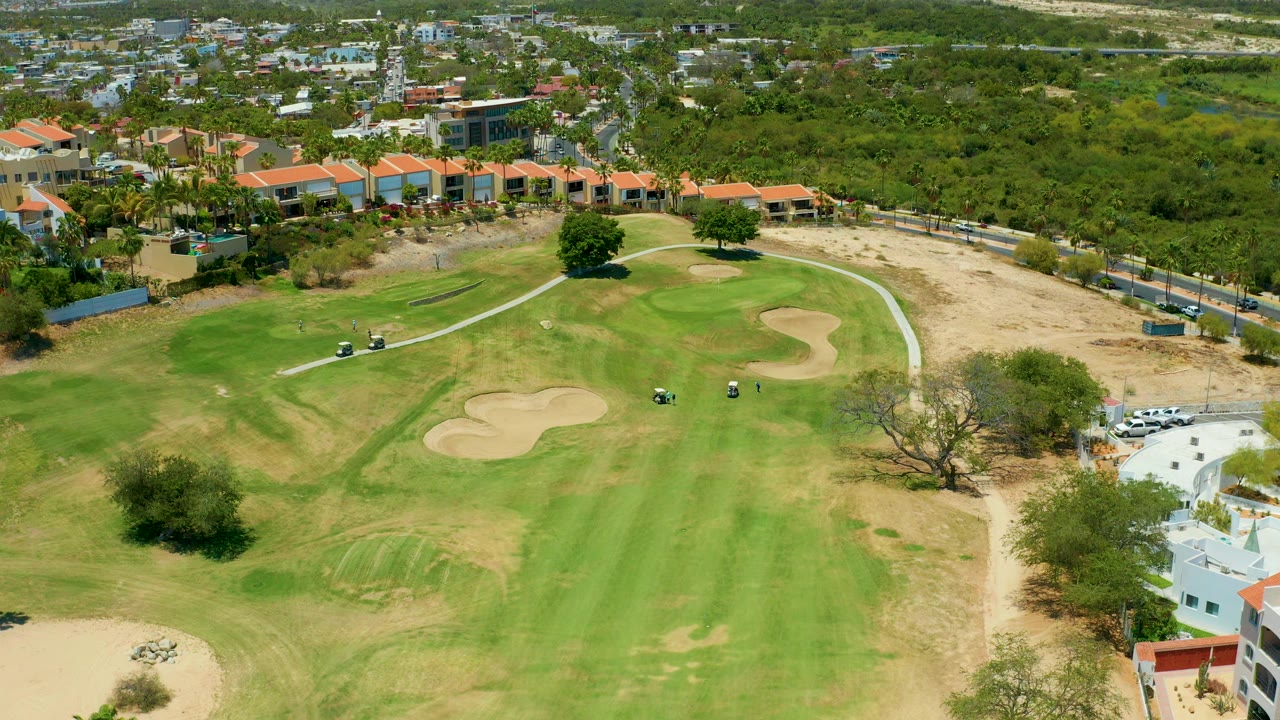 Aerial frame over a San Jose Del Cabo golf course showing green fairway, pale sand traps, a curving cart path, surrounding trees, nearby buildings, and no visible people.