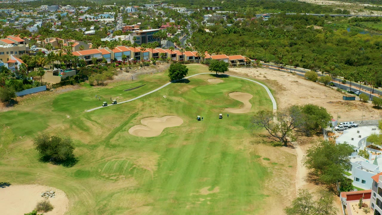 One second into the San Jose Del Cabo aerial, the fairway fills the lower frame while carts sit near the curving path, bunkers sit left and right of the green, and no people are visible.
