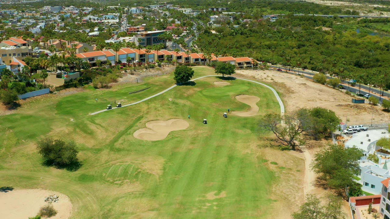 Aerial frame from San Jose del Cabo showing green fairways, tan sand traps, a pale cart path, nearby buildings, trees, and no visible people at the start of the clip.