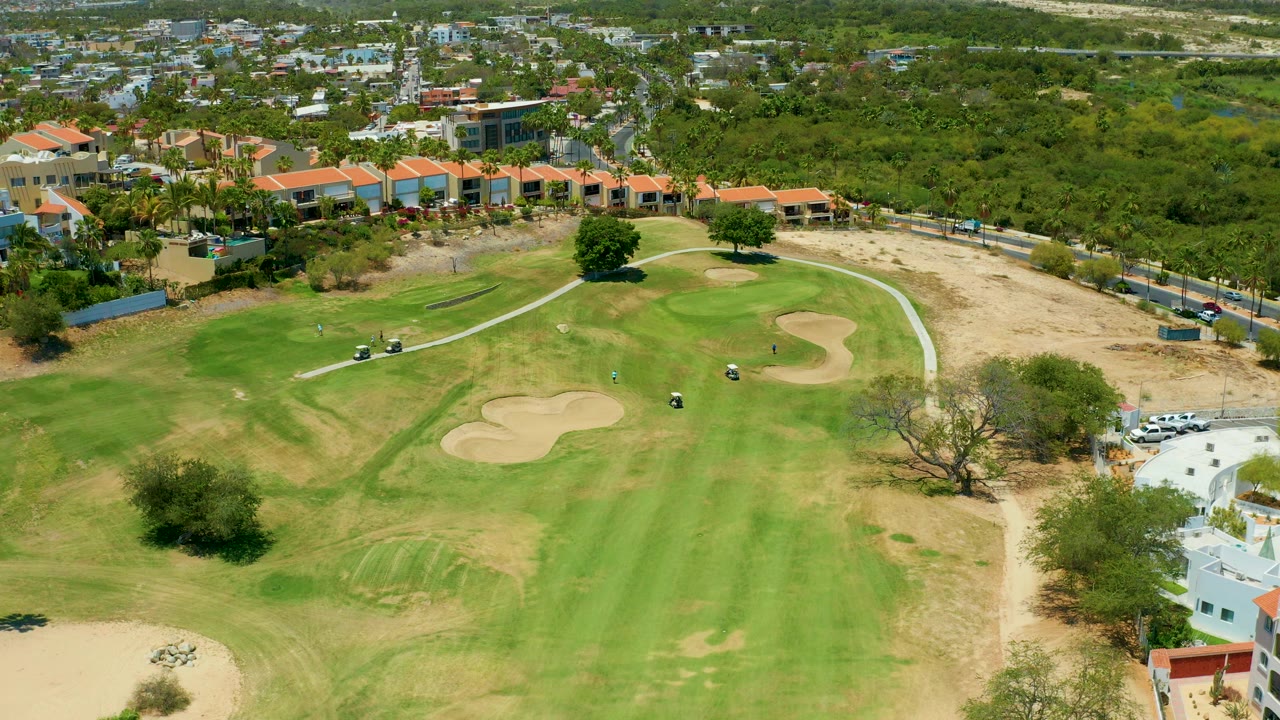 Aerial frame over a San Jose del Cabo golf course with green fairways, pale sand traps, small golf carts, orange-roofed buildings, dry ground, and no visible people.