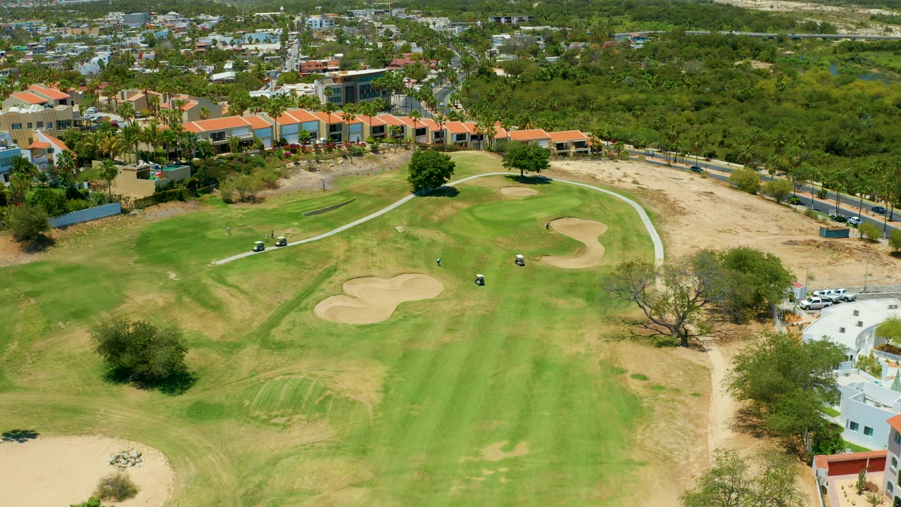 Drone frame in San Jose del Cabo showing fairway stripes, multiple sand traps, a curved cart path, sparse trees, roadside vehicles, and no visible people.