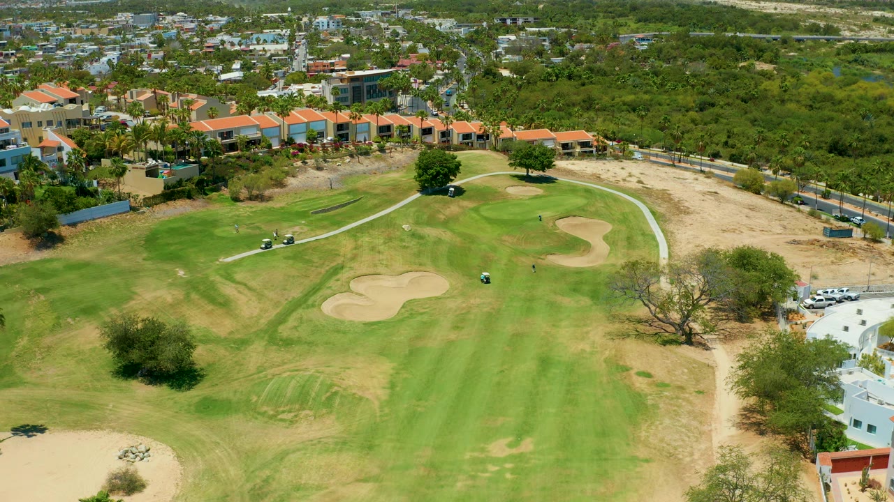 Aerial view in San Jose del Cabo with the fairway centered, two tan sand traps near the green, a cart path curving left, background buildings, and no visible people.