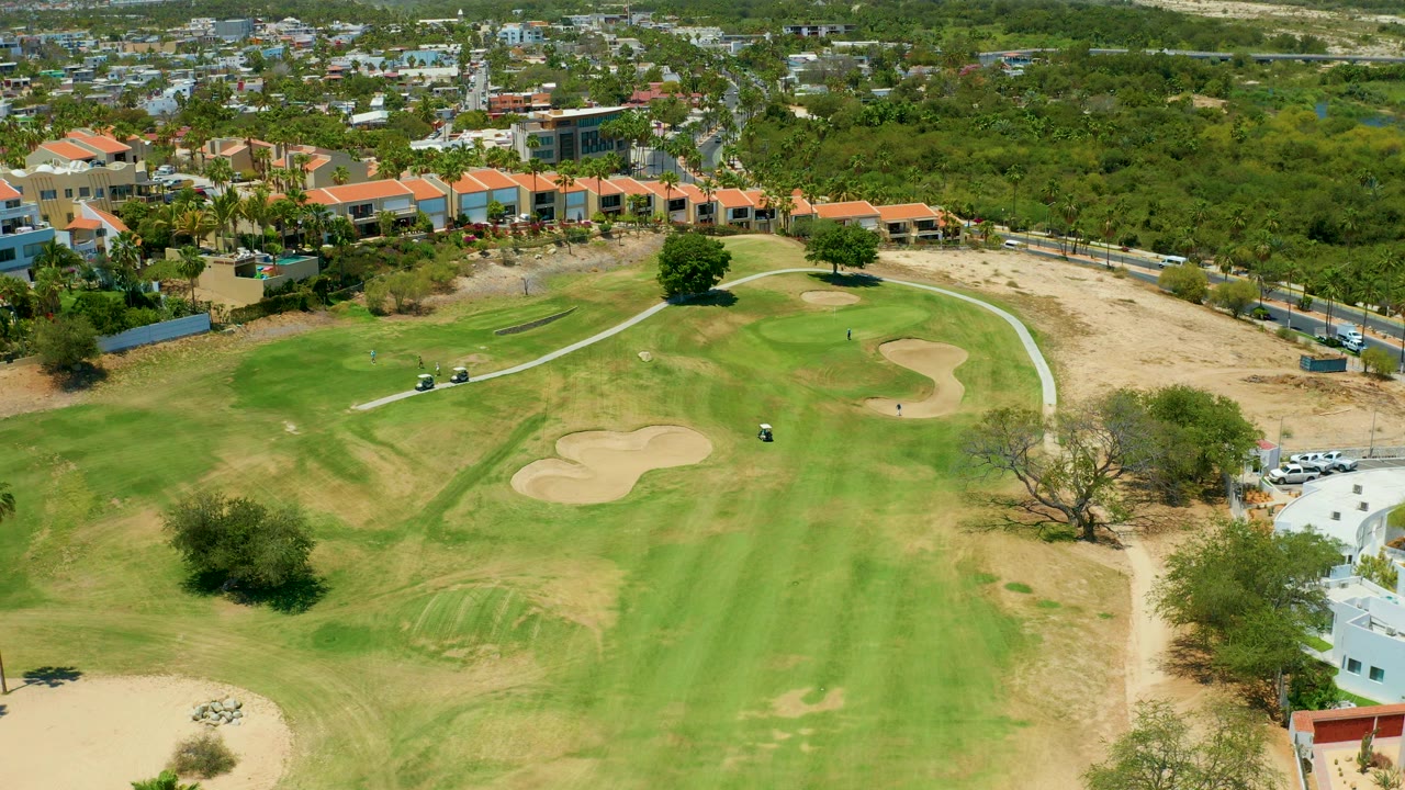 Aerial frame from San Jose del Cabo showing a golf course fairway, tan bunkers, a curving cart path, dry rough, trees, and low buildings beyond the course, with no clearly visible people.