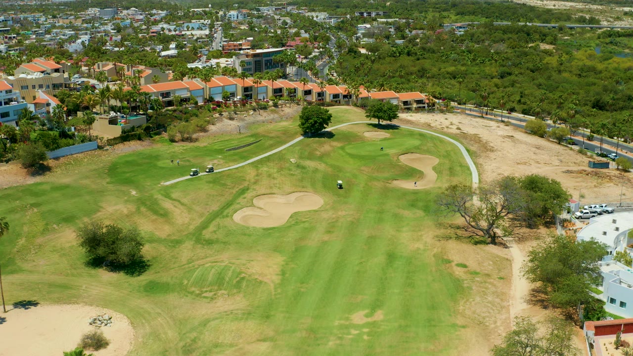 San Jose del Cabo golf fairway seen from above, with pale sand traps near center, small golf carts on the path, dry edges, and residential buildings beyond the green, with no visible people.