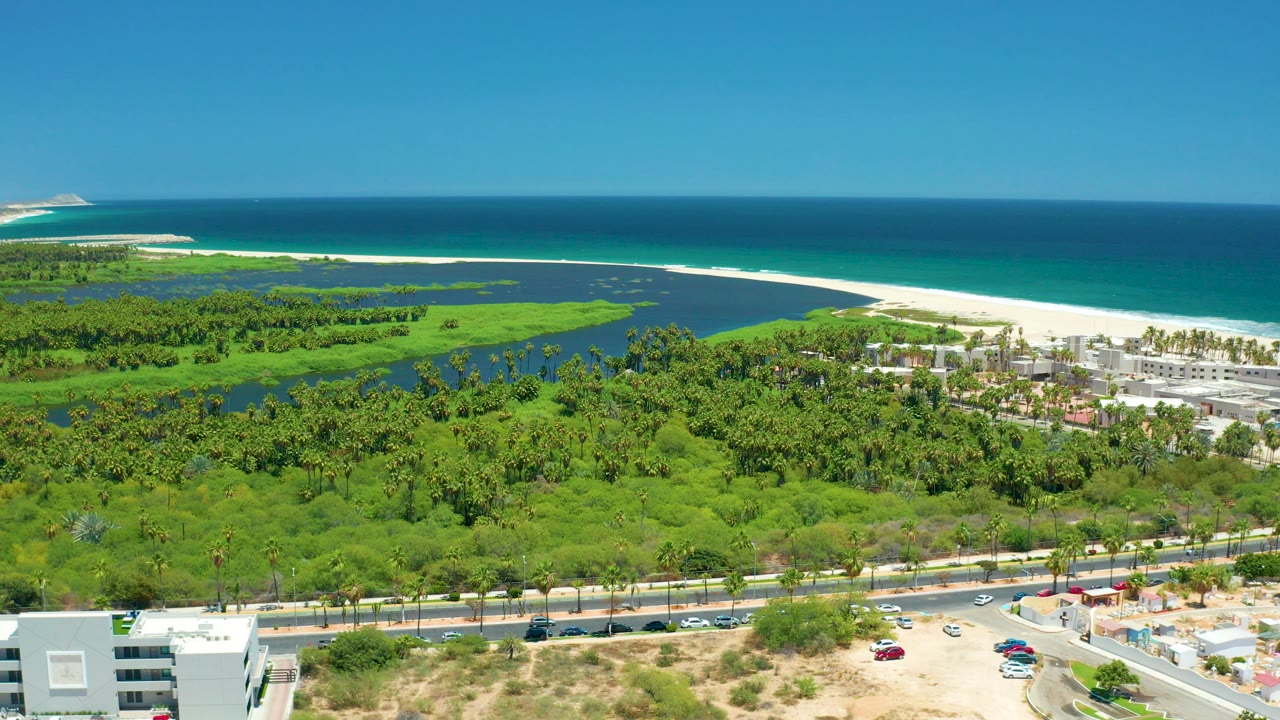 Opening aerial frame over San Jose Del Cabo showing a coastal road with cars, dense green vegetation, wetlands, white beach, blue ocean, and no visible people.