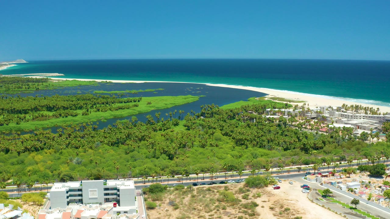 Opening aerial frame over San Jose Del Cabo showing foreground buildings and road, green lagoon vegetation, a white beach, blue ocean, and no visible people.