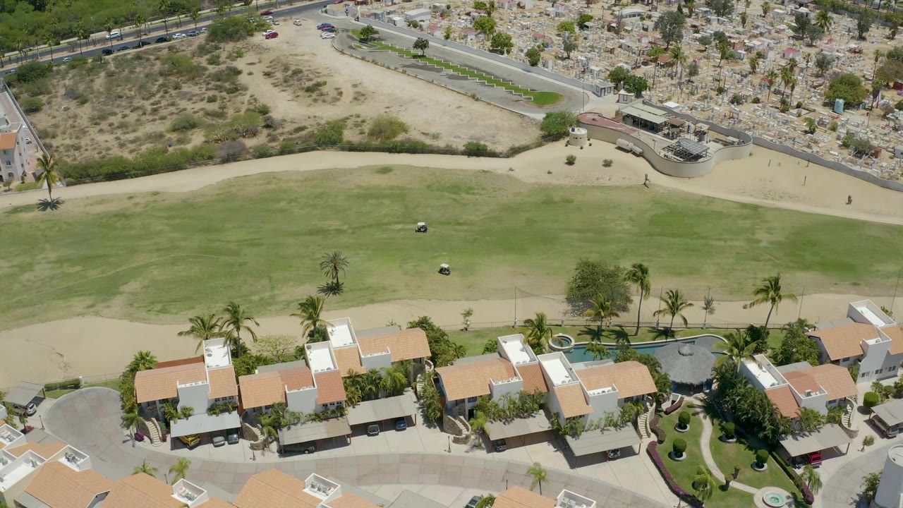 High aerial view in San Jose del Cabo with the golf fairway centered, brown-roofed residential buildings below, roadside traffic above, and no visible people.