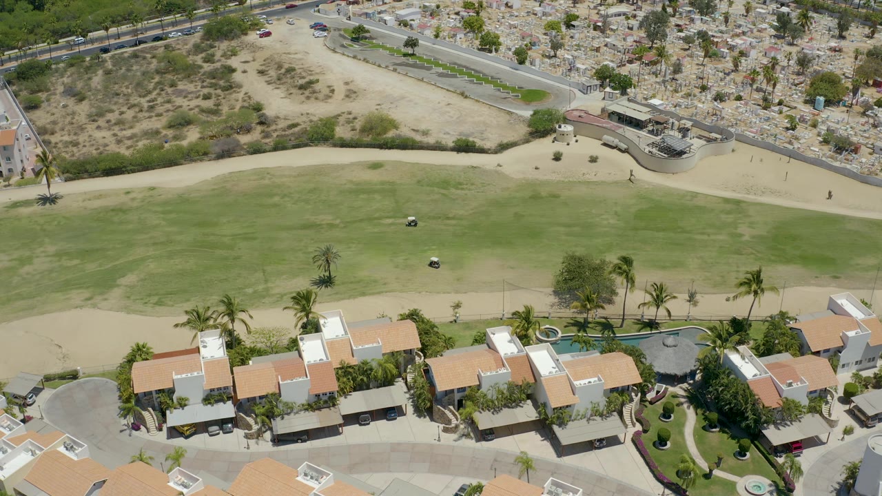 Drone view over San Jose del Cabo residential roofs and palms, with golf-course grass in the middle, dry land and a road in the background, and no visible people.