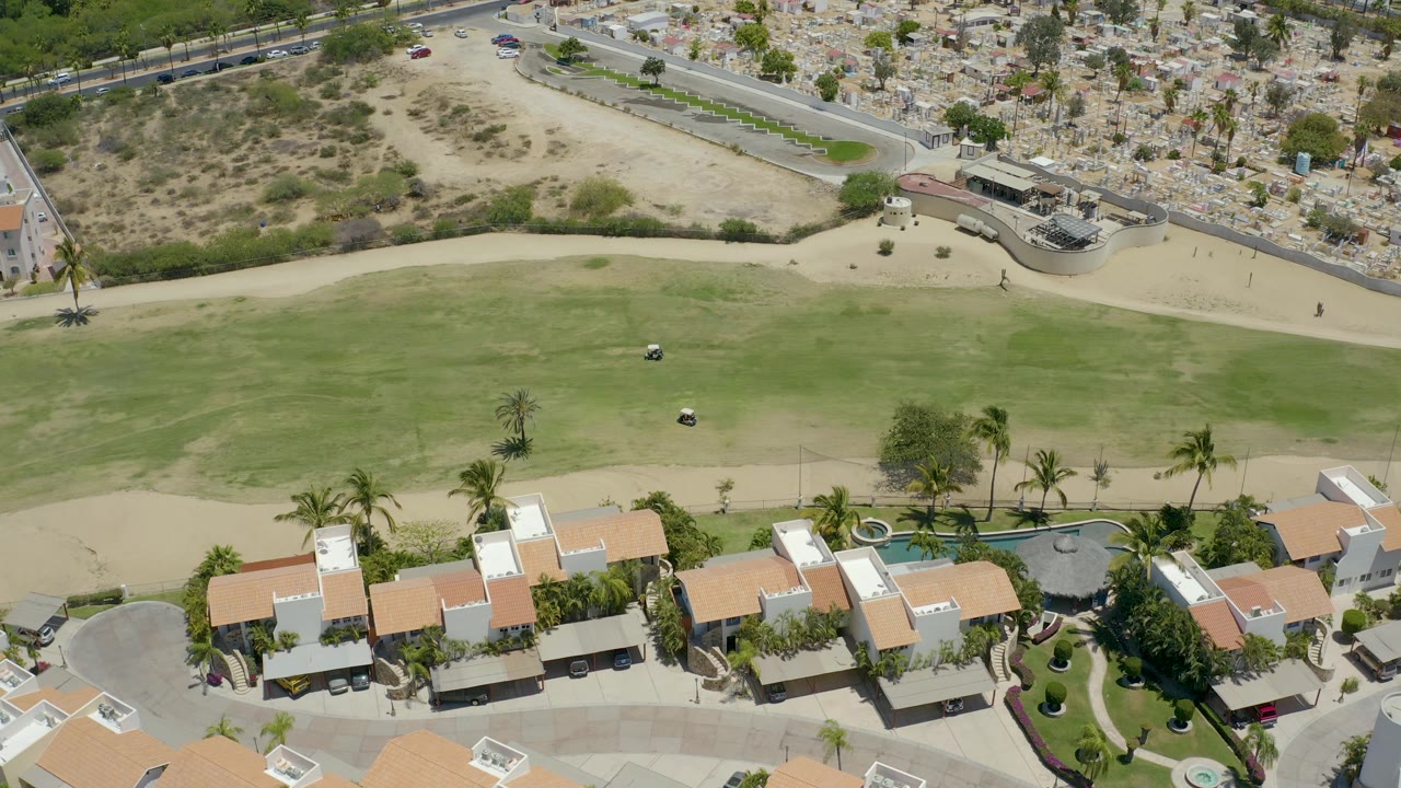Aerial frame of homes, golf course, palm trees, and a road crossing dry terrain.