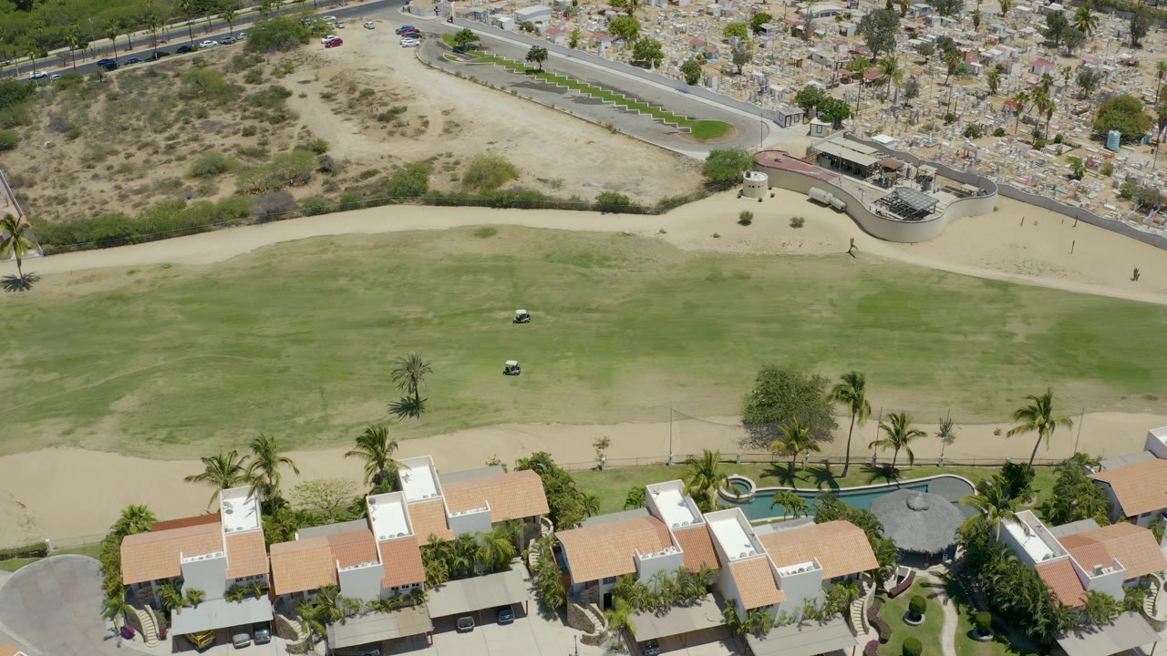 A high drone frame over San Jose Del Cabo shows the golf cart on the open grass, resort rooftops and a pool along the bottom edge, sandy terrain above, and no visible people.