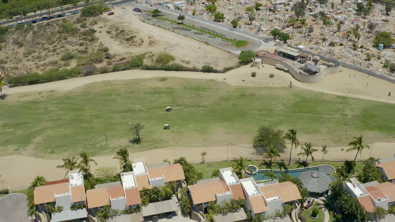 San Jose Del Cabo golf course seen from above with a small cart on the fairway, palm trees near red-roofed buildings, sandy roads and hillside housing beyond, and no visible people.