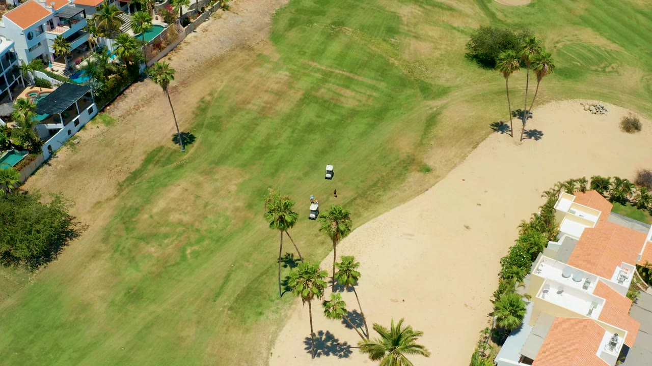 A high-angle aerial frame over a San Jose Del Cabo golf course shows two small white carts on the fairway, a large sand area, palm trees, and nearby homes, with no visible people.