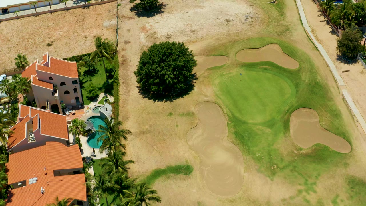 Overhead frame in San Jose del Cabo shows red-roofed villas, a curved blue pool, palms, dry grass, a golf green, sand traps, and no visible people.