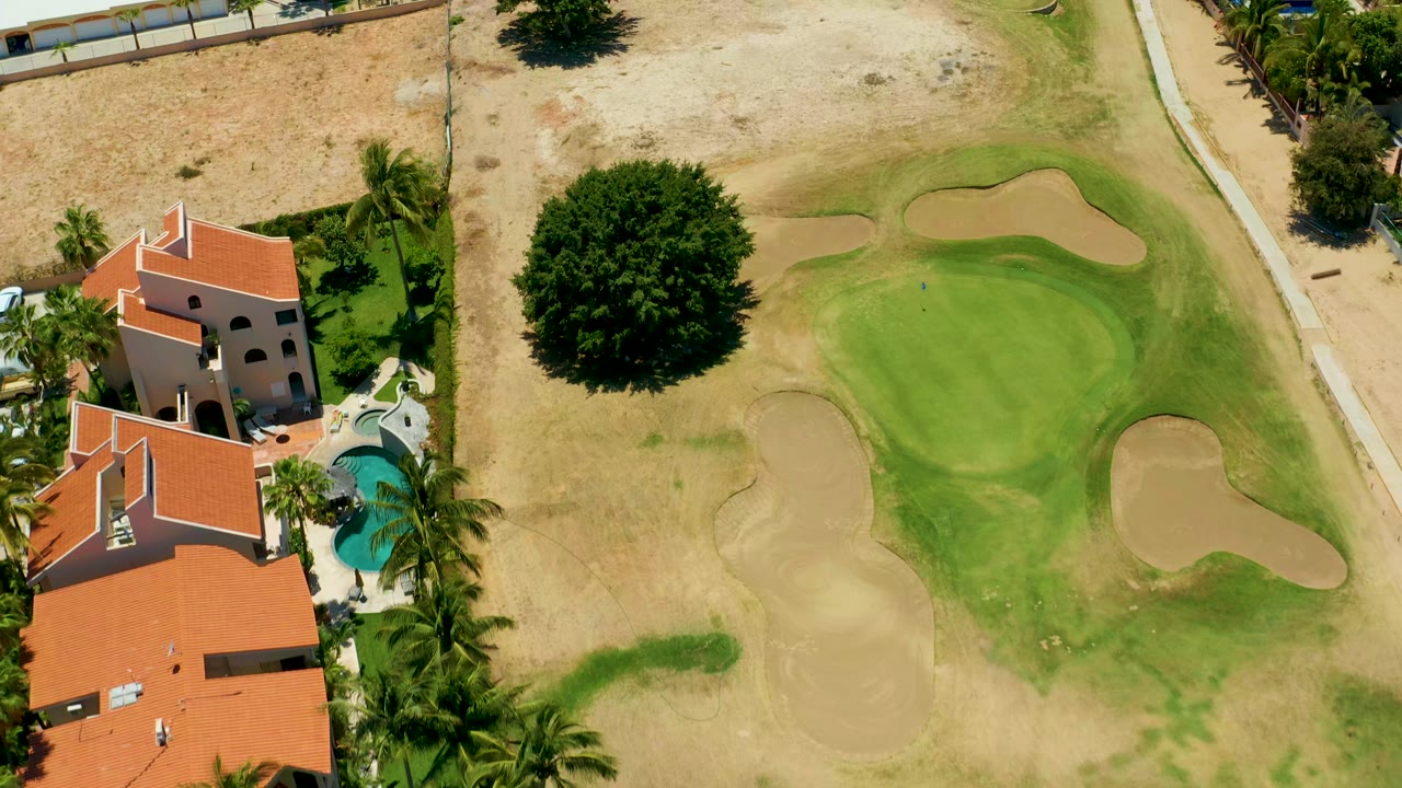 San Jose del Cabo aerial frame shows the golf green and bunkers taking more of the view while red-roofed homes and a pool remain along the left edge, with no visible people.