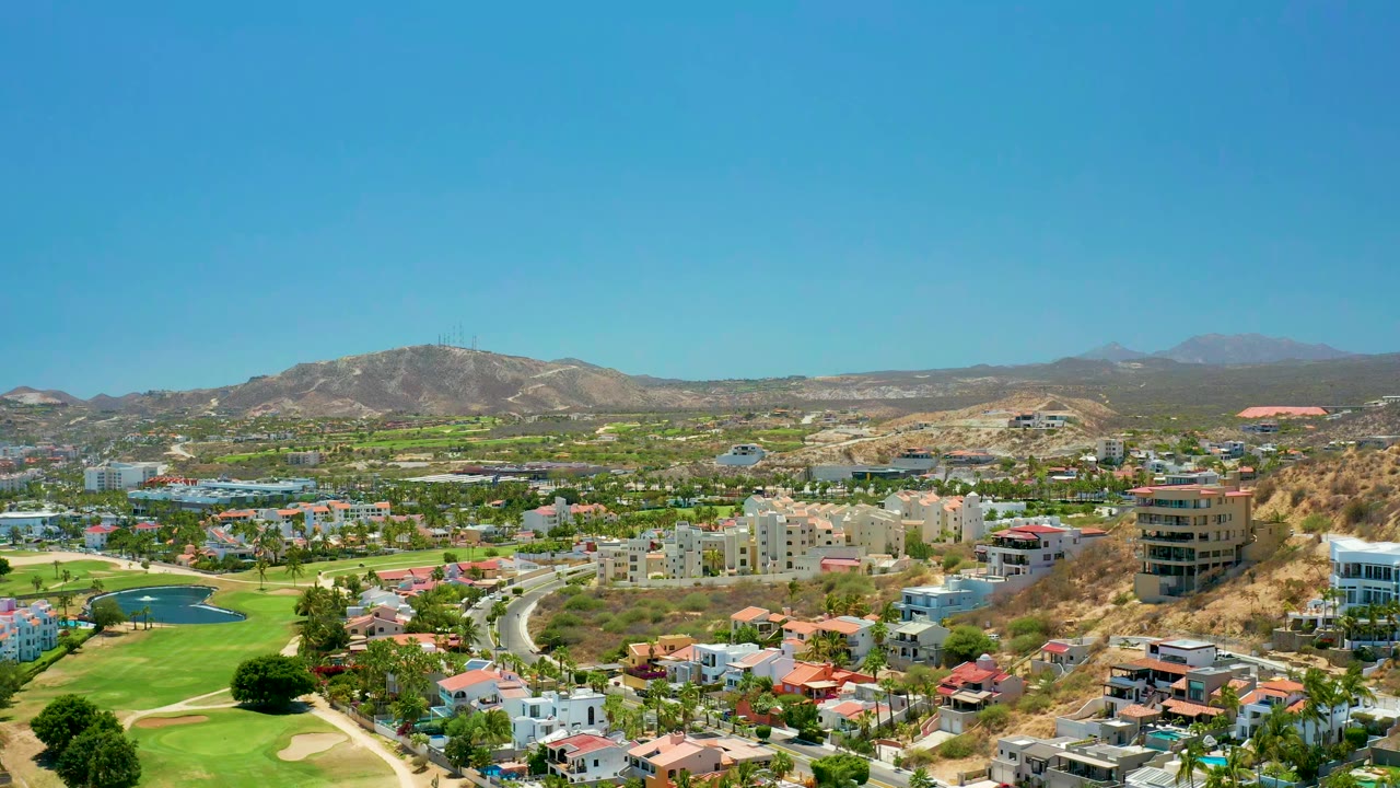 Opening frame over San Jose del Cabo shows a curved road beside green fairways, a small pond, hillside homes, distant mountains, and no visible people.