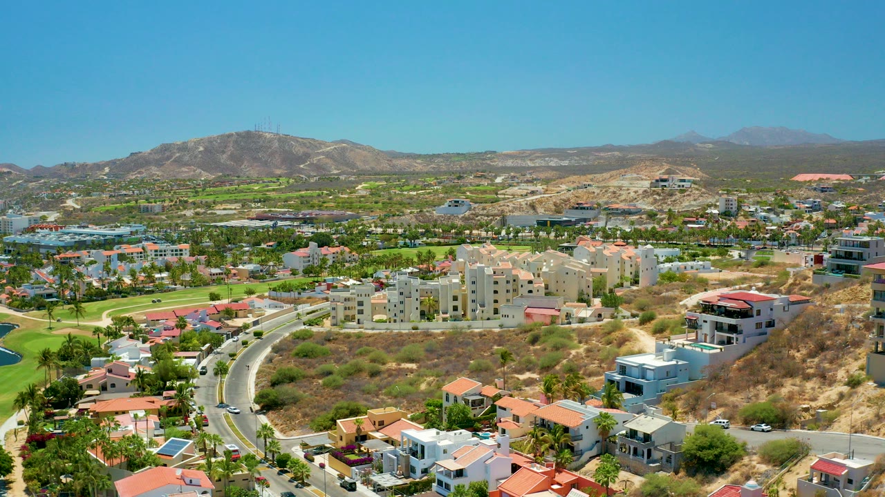 Wide aerial frame over San Jose Del Cabo showing a curved residential road, hillside houses, green landscaping, distant mountains, clear blue sky, and no visible people.