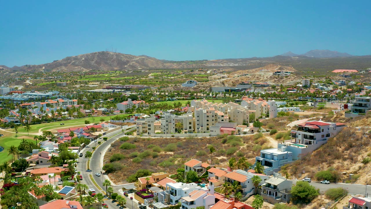 Wide 4K aerial frame of San Jose Del Cabo residential development, with a winding road, red-roofed homes, pale apartment buildings, tan hillside terrain, distant mountains, and no visible people.