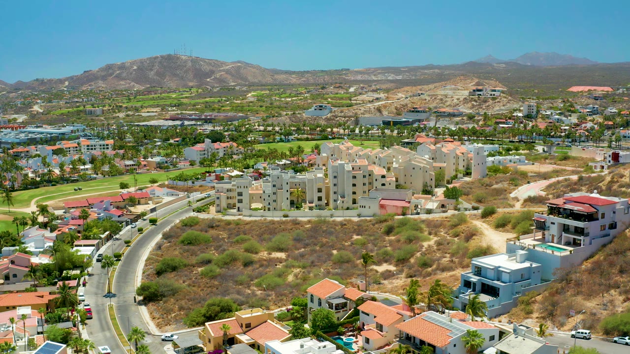 Aerial frame in San Jose del Cabo showing the road bending through foreground homes, beige apartment buildings in the center, green patches behind them, distant hills, and no visible people.