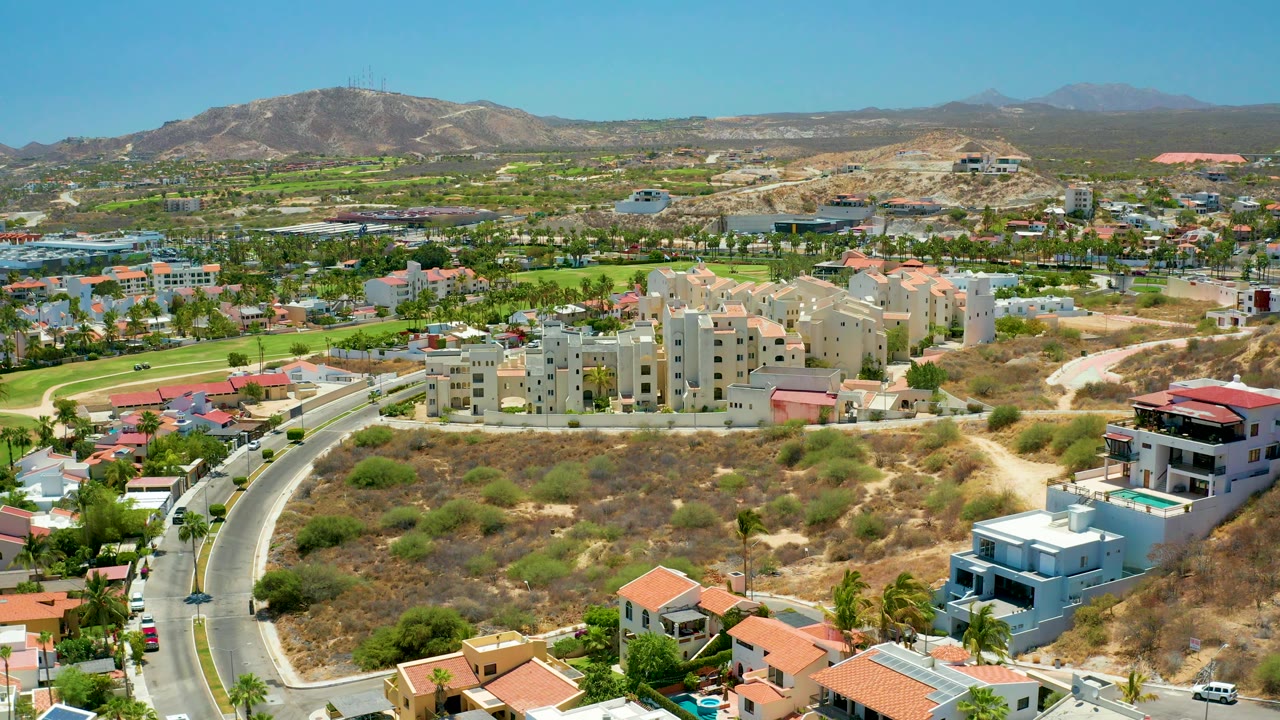 Aerial neighborhood view with beige buildings, curved road, dry terrain, and hills.