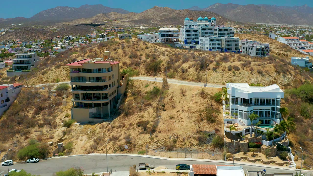 Aerial frame over San Jose Del Cabo showing a dry hillside with modern homes, a curved road at the bottom, distant mountains, and no visible people.