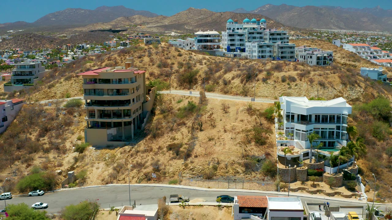 Aerial view in San Jose Del Cabo with a beige multi-story home near center, a white modern house to the right, dry brush on the slope, a lower road with vehicles, and no visible people.
