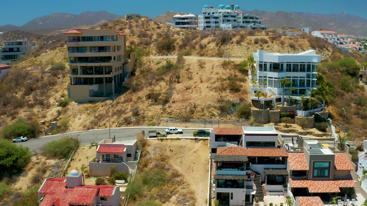 Opening aerial frame over San Jose Del Cabo hillside residences, with red-roofed homes in the foreground, a curved road below the slope, modern buildings on the hill, distant mountains, and no visible people.