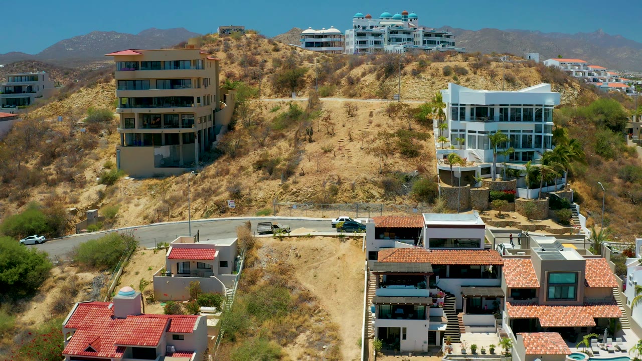 Aerial view in San Jose Del Cabo with foreground rooftops lower in frame, cars along the curved road, dry brush on the slope, modern hillside residences, mountains, and no visible people.