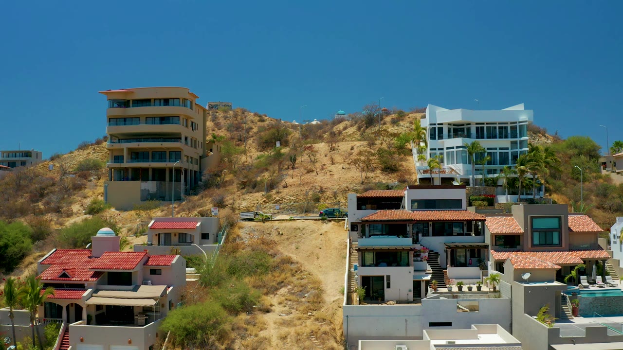 A dry sandy cut through the hillside separates modern homes in San Jose Del Cabo, with sparse vegetation, bright midday light, and no visible people.