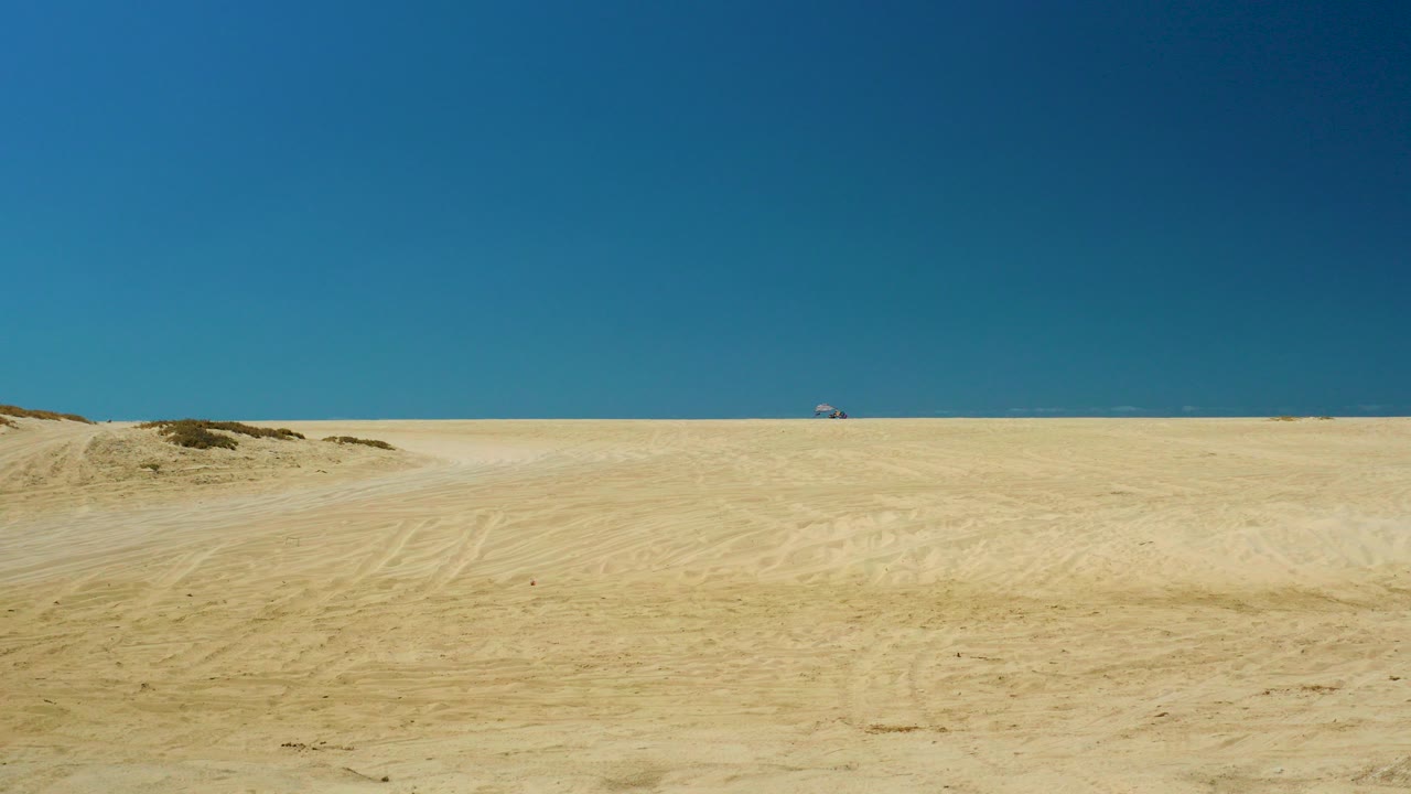 Wide La Laguna Beach Los Cabos sandy desert view under a deep blue sky, with low dune mounds, tire marks, a distant sea horizon, and no visible people.