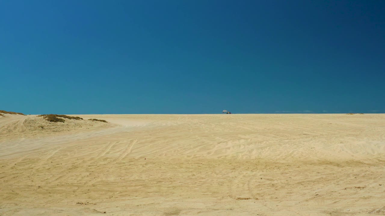 La Laguna Beach Los Cabos sand flats stretch toward a thin ocean line, with a small distant vehicle near the horizon and no visible people.