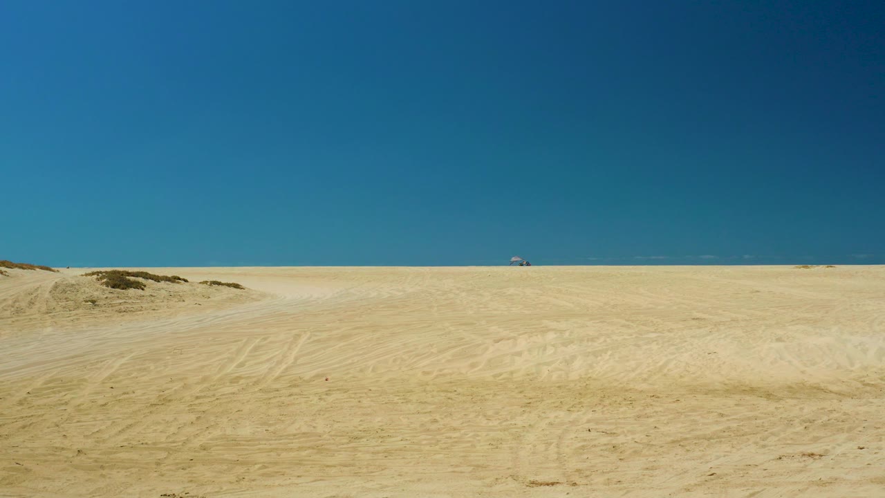 A broad La Laguna Beach Los Cabos sandy landscape shows faint vehicle tracks, scrub-topped dune rises, a distant ocean horizon, and no visible people.