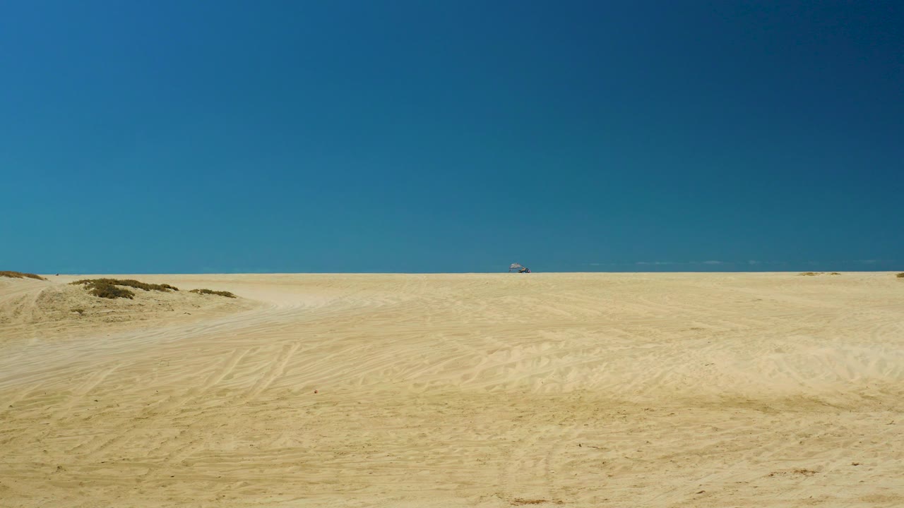 A low sandy mound sits in the foreground of the La Laguna Beach Los Cabos desert scene, with open sand, a flat sea horizon, and no visible people.