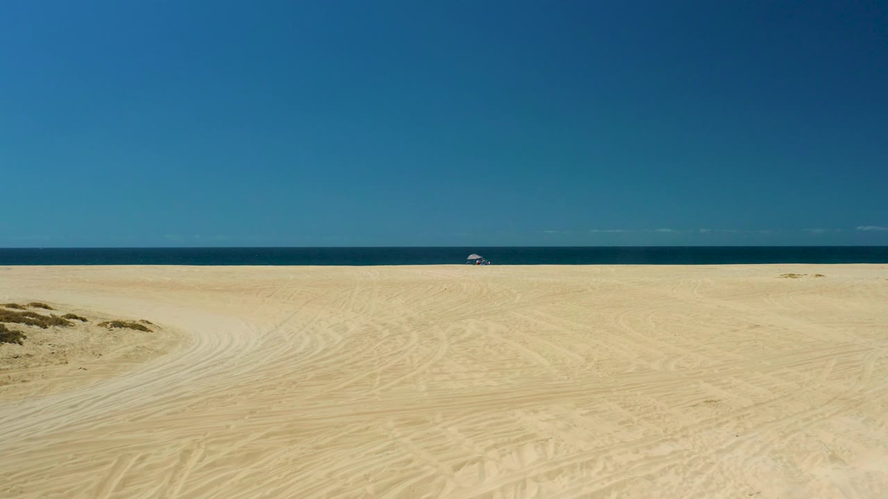 At 0:02, the La Laguna Beach Los Cabos shoreline is seen from a wide elevated angle with broad sand, faint vehicle tracks, small dune mounds, deep blue ocean, and a tiny umbrella with a person in the distance.