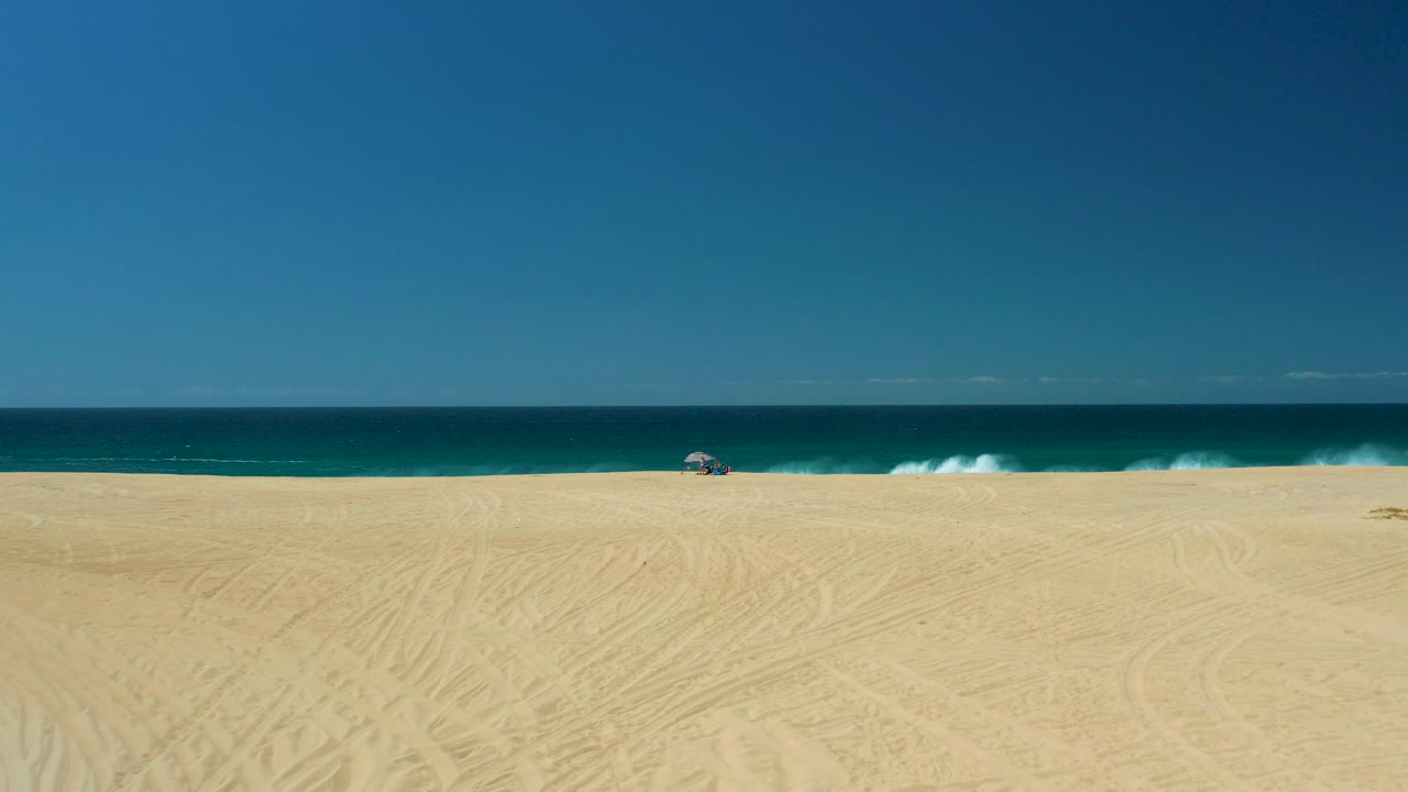 Wide elevated view of La Laguna Beach in Los Cabos, Mexico, with broad sand in front, blue ocean beyond, a small umbrella near the waterline, and no visible people.